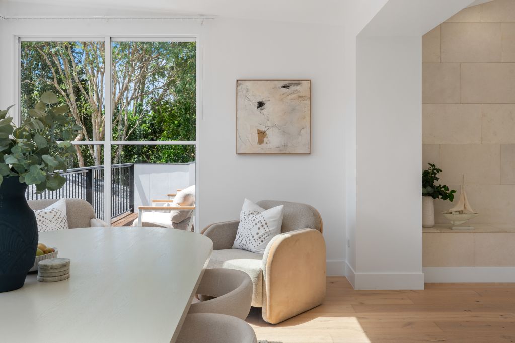 Bright minimalist living room with white walls, large window, beige chair, dining table, and a plant.