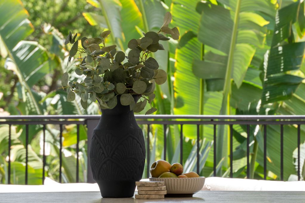 Outdoor table with a black vase of flowers, a fruit bowl, and books, against lush green banana leaves.