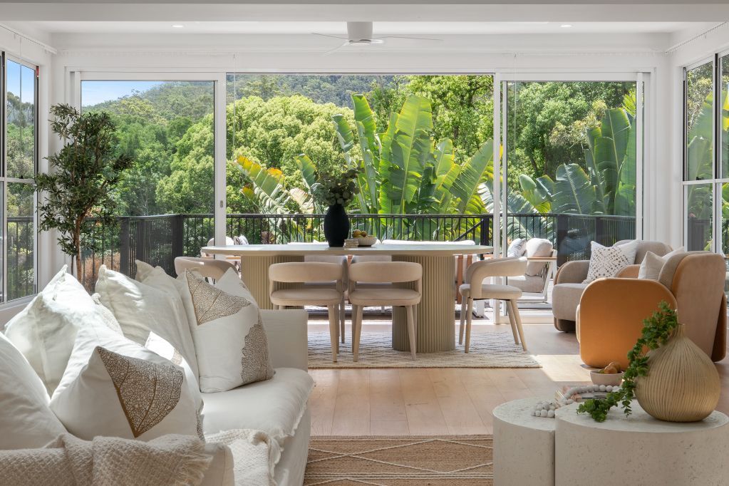 Bright living room with white sofas, wooden dining table, and open glass walls overlooking lush greenery