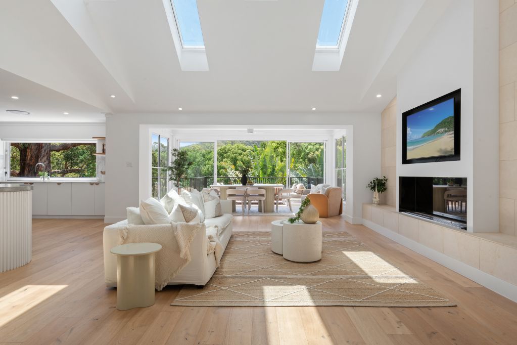Bright modern living room with white sofas, skylights, fireplace, and large windows overlooking greenery