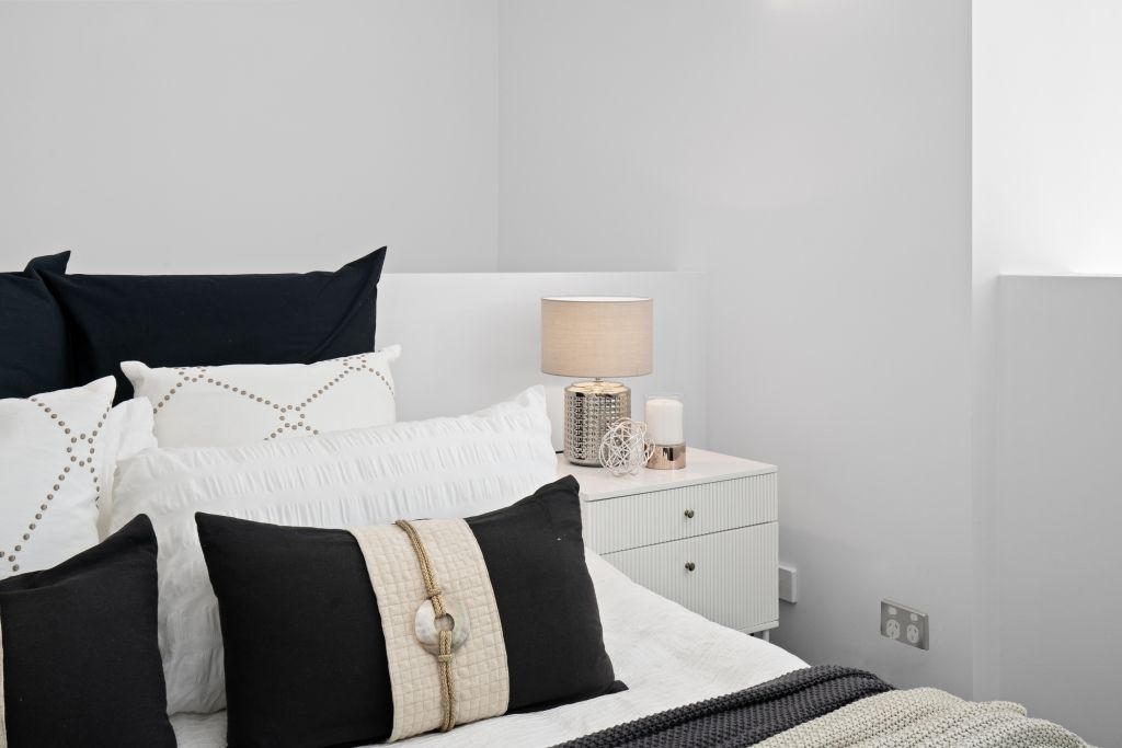 A bedroom with a bed , nightstand , lamp and pillows.