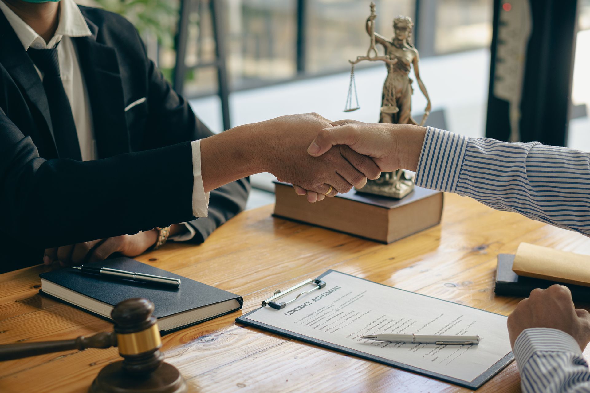 Two people shaking hands over a desk, with a gavel, law books, and Lady Justice statue.