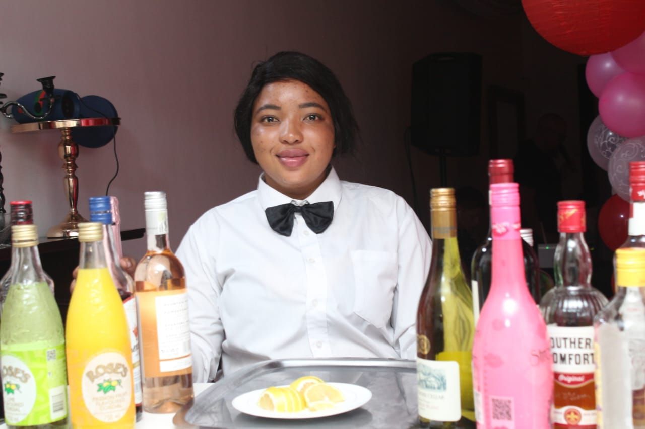 A woman in a bow tie stands in front of a table full of bottles of alcohol