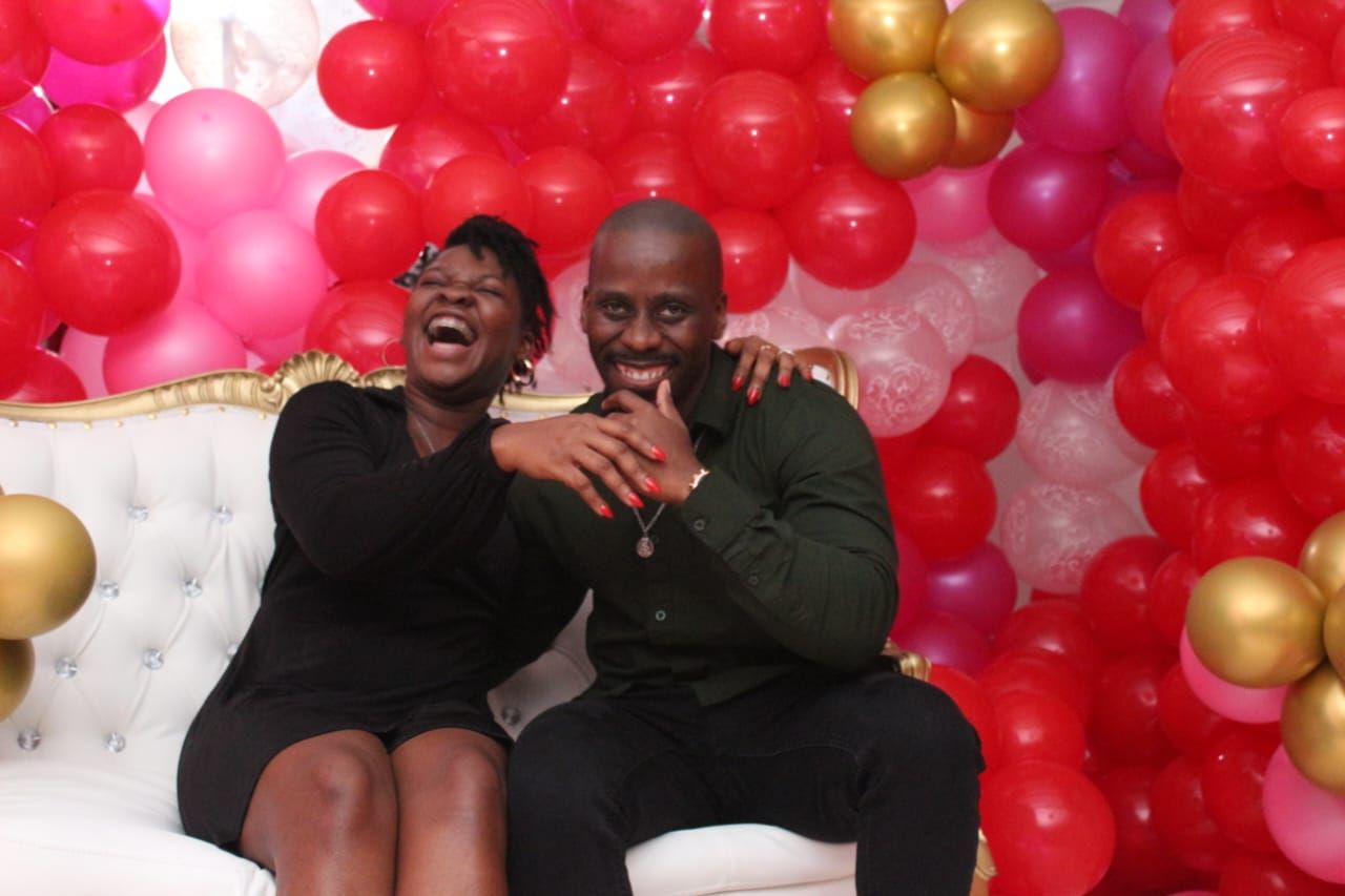 A man and a woman are sitting on a couch in front of balloons.