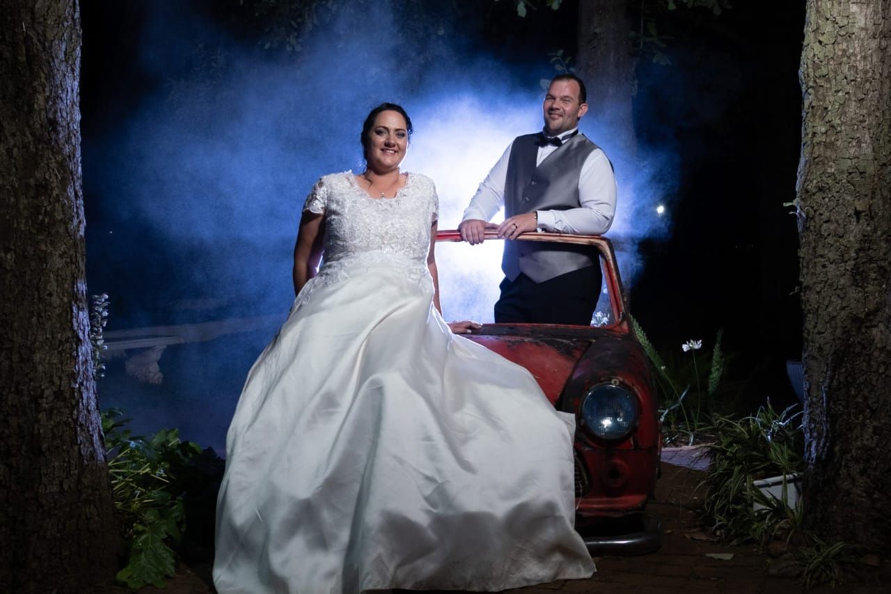 A bride and groom are posing for a picture in front of a red car.