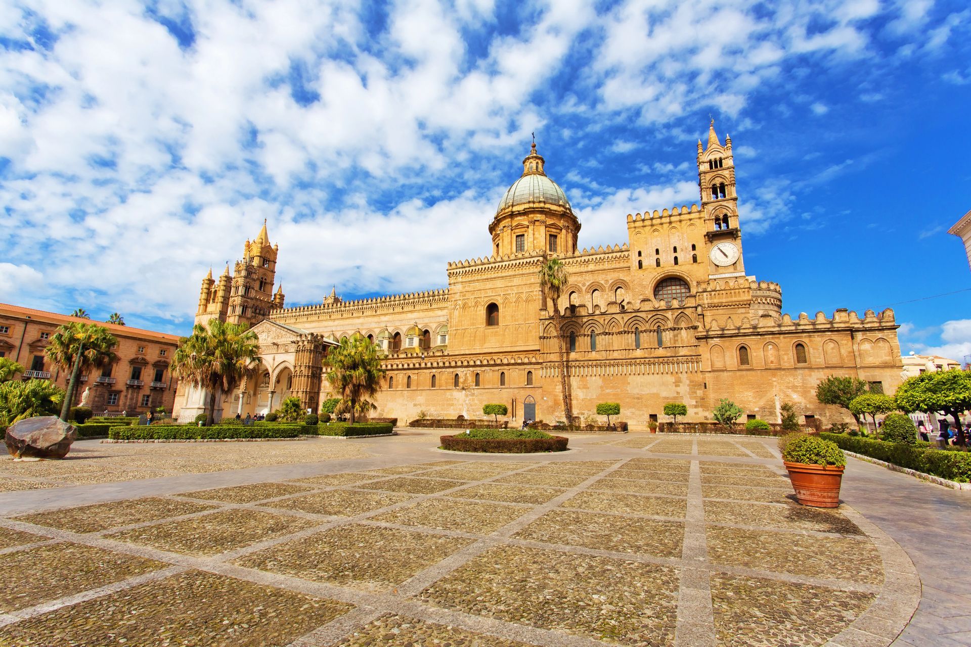 Il Duomo di Palermo, in Sicilia, è uno storico edificio in pietra dorata con una maestosa cupola e torri, sotto un cielo azzurro e nuvoloso.