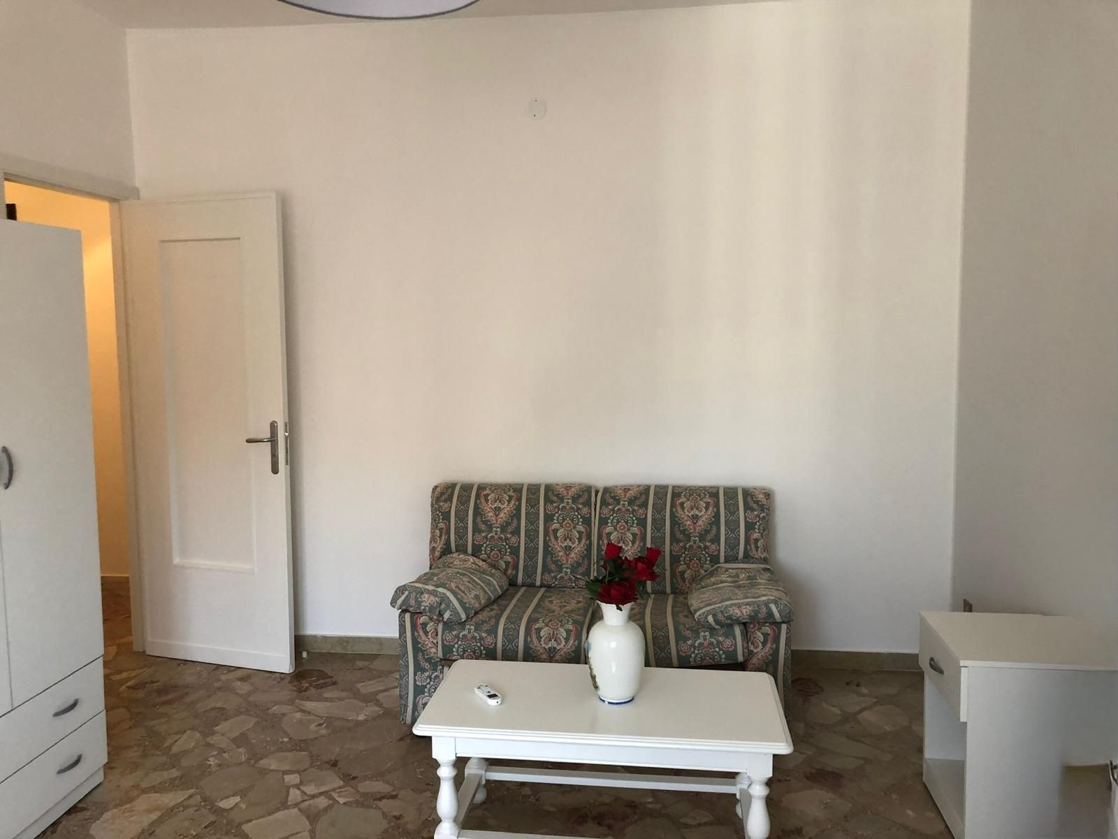 Un divano a fantasia è addossato a una parete bianca, con un tavolino da caffè bianco, un vaso di fiori rossi e una porta sullo sfondo.