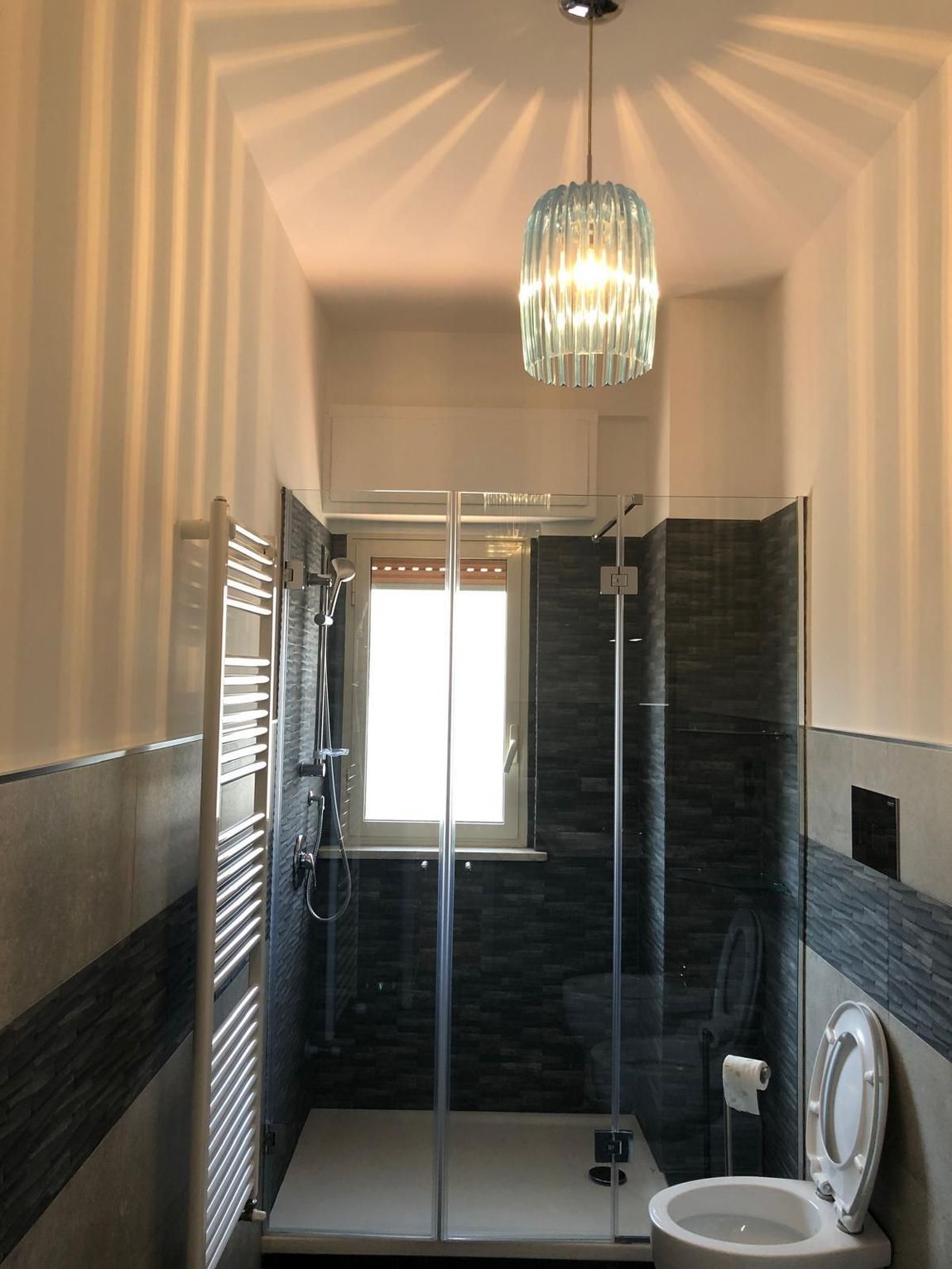 Un bagno moderno con doccia con box in vetro, pareti rivestite con piastrelle scure, un portasciugamani a scala bianco e una lampada a sospensione dal design unico.