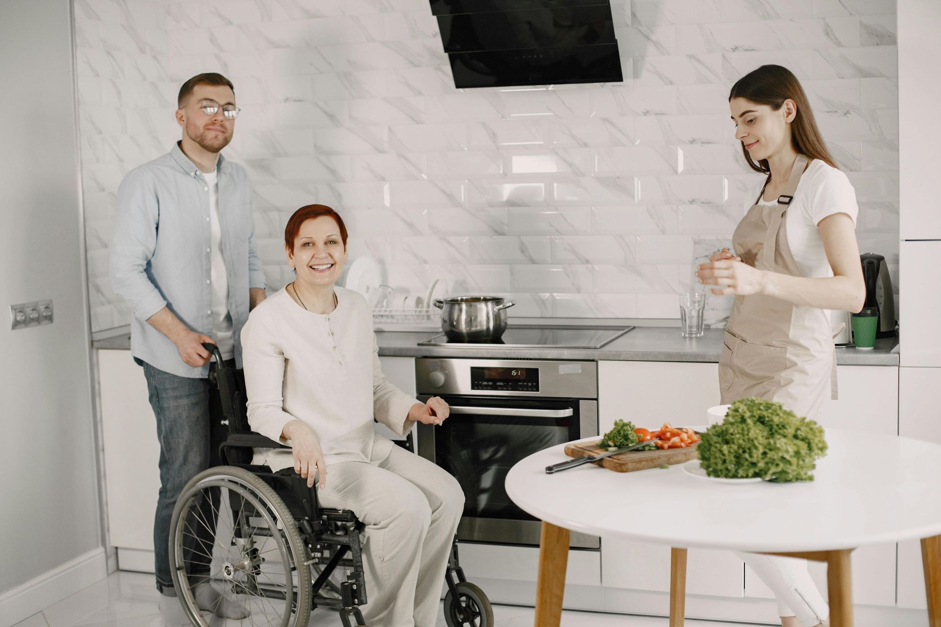 NDIS provider assisting a woman in a wheelchair in a kitchen while another caregiver prepares food.
