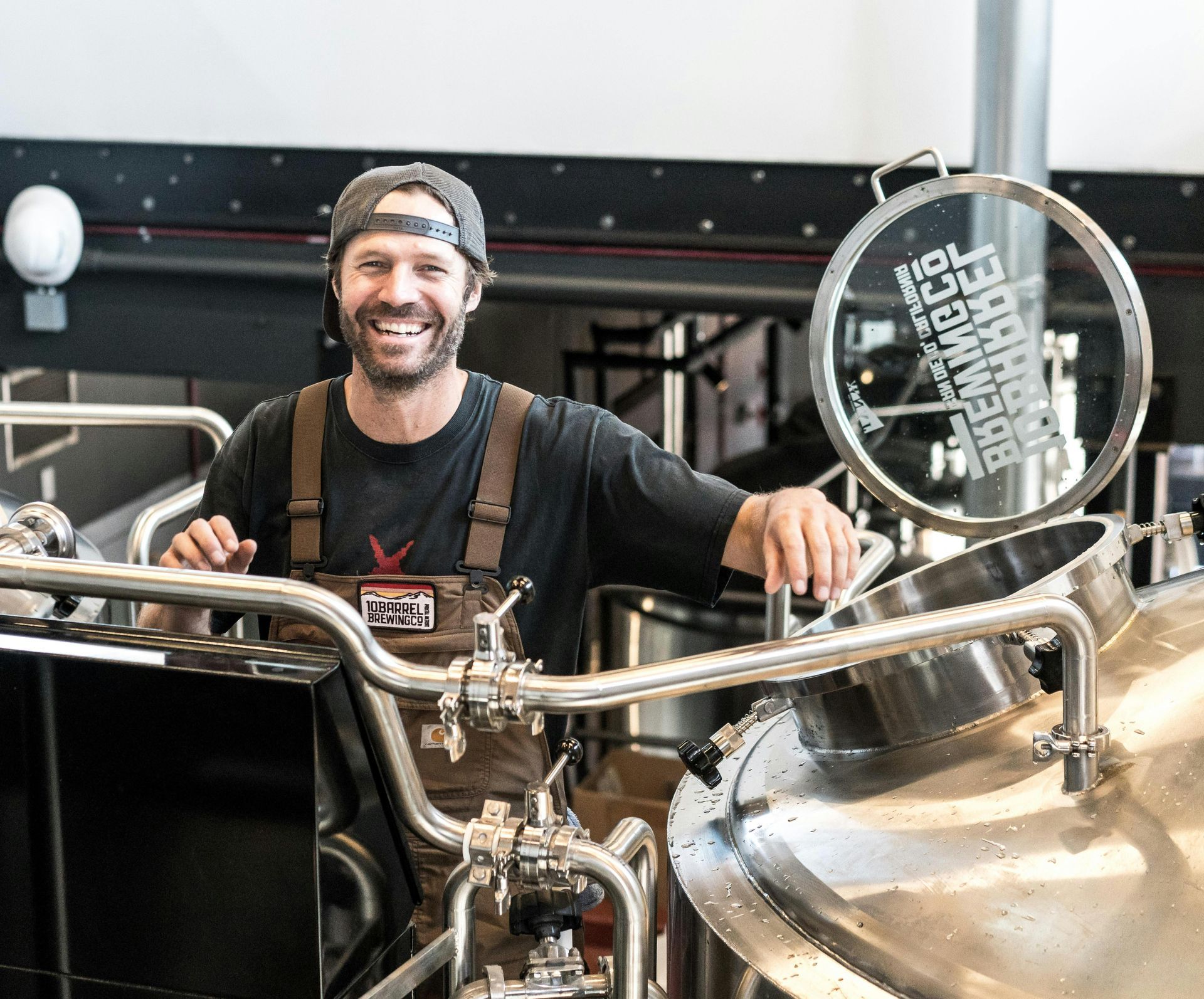 Man in overalls smiles, standing at stainless steel brewing equipment in a brewery.