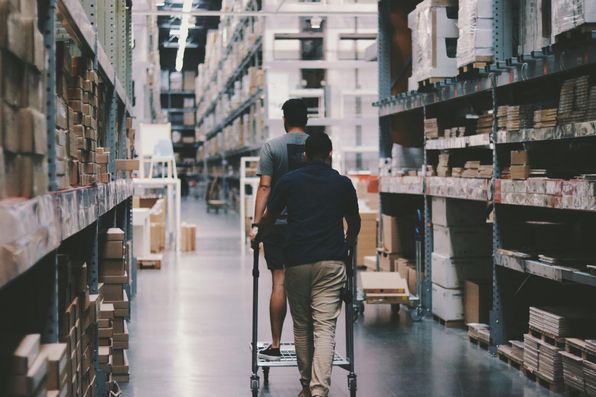 Two people walk down an aisle in a warehouse. One is pushing a cart. Shelves are stocked with boxes.