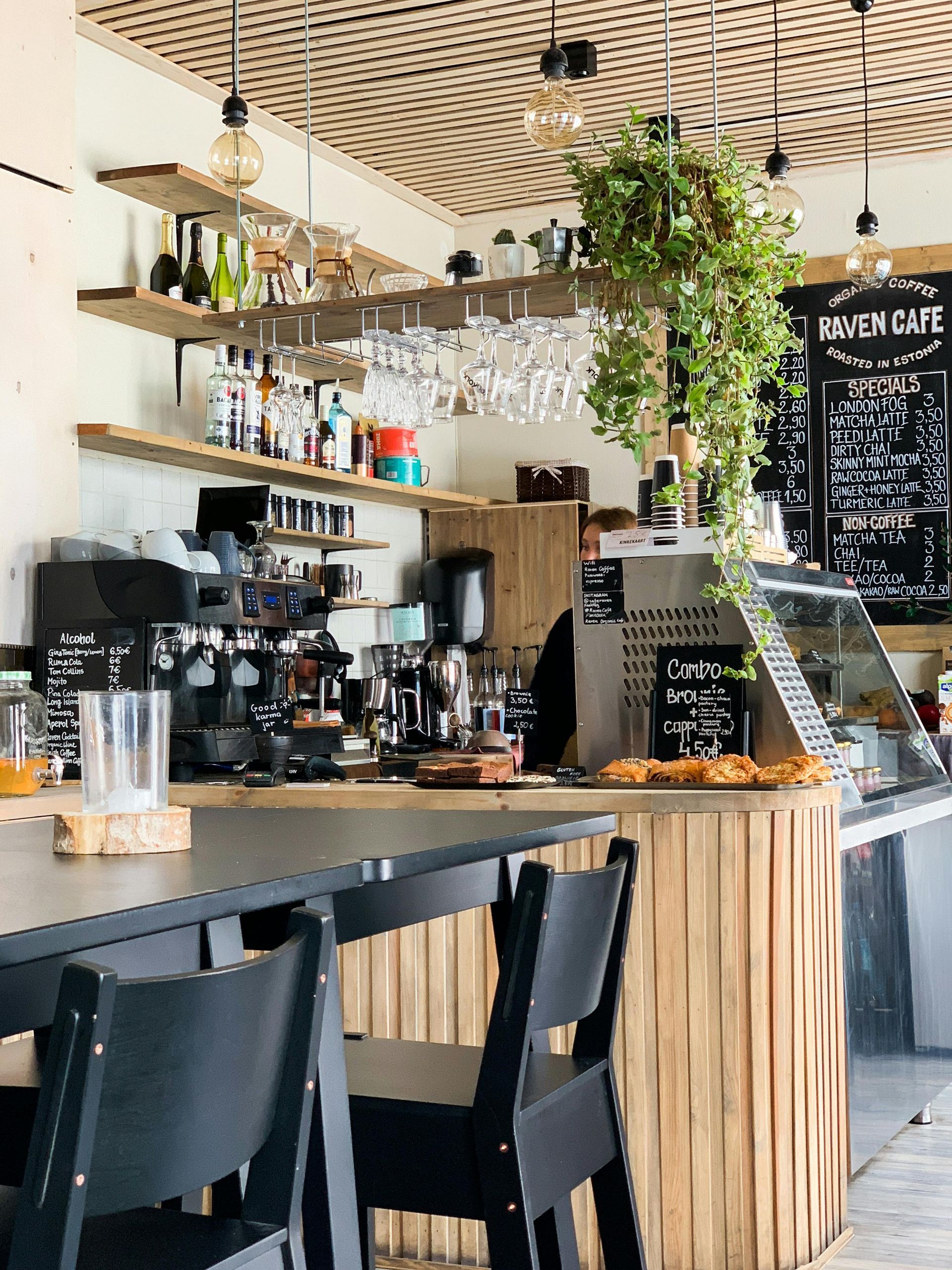 Cafe interior: bar with espresso machine, shelving with bottles, hanging plants, blackboard menu.