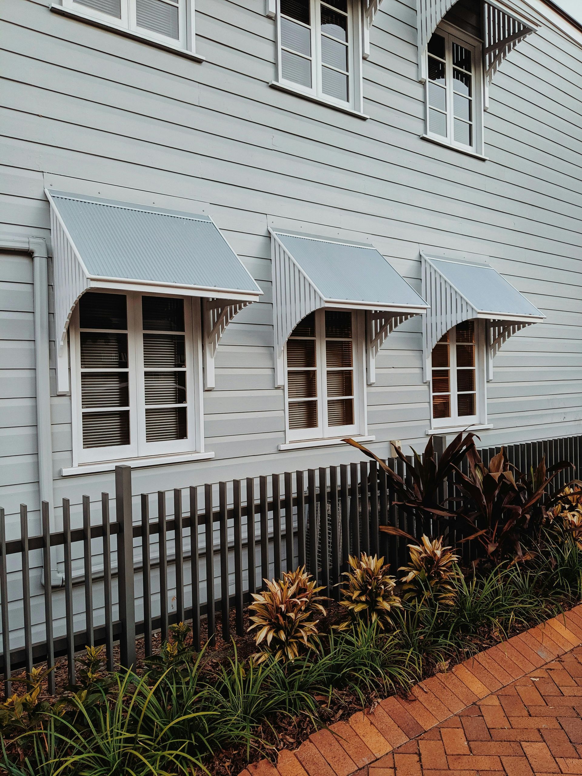 Grey wooden house with white windows, awnings, and a brick path, behind a wooden fence with plants.