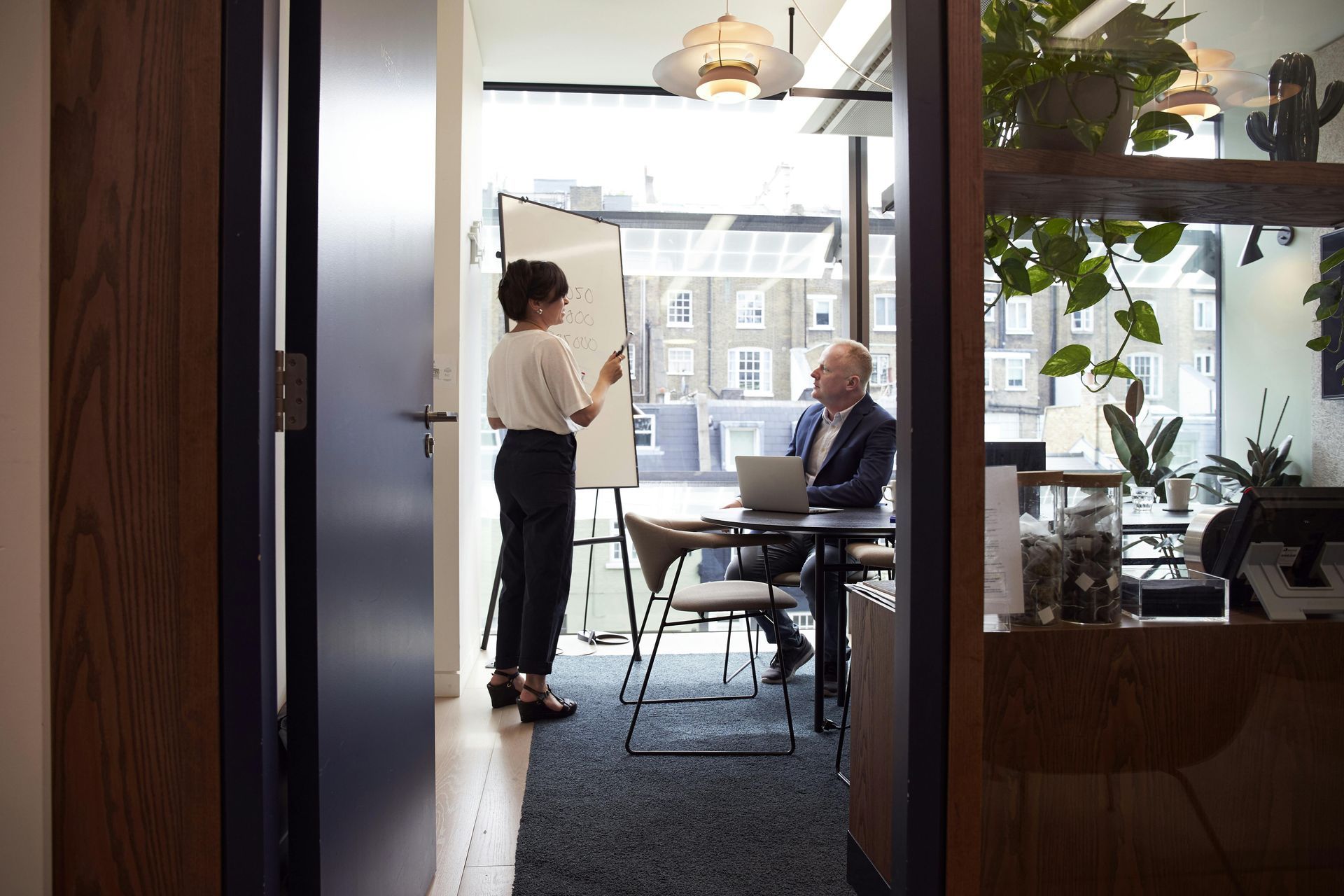 Woman presenting to a man in a modern office; whiteboard, laptop, natural light.