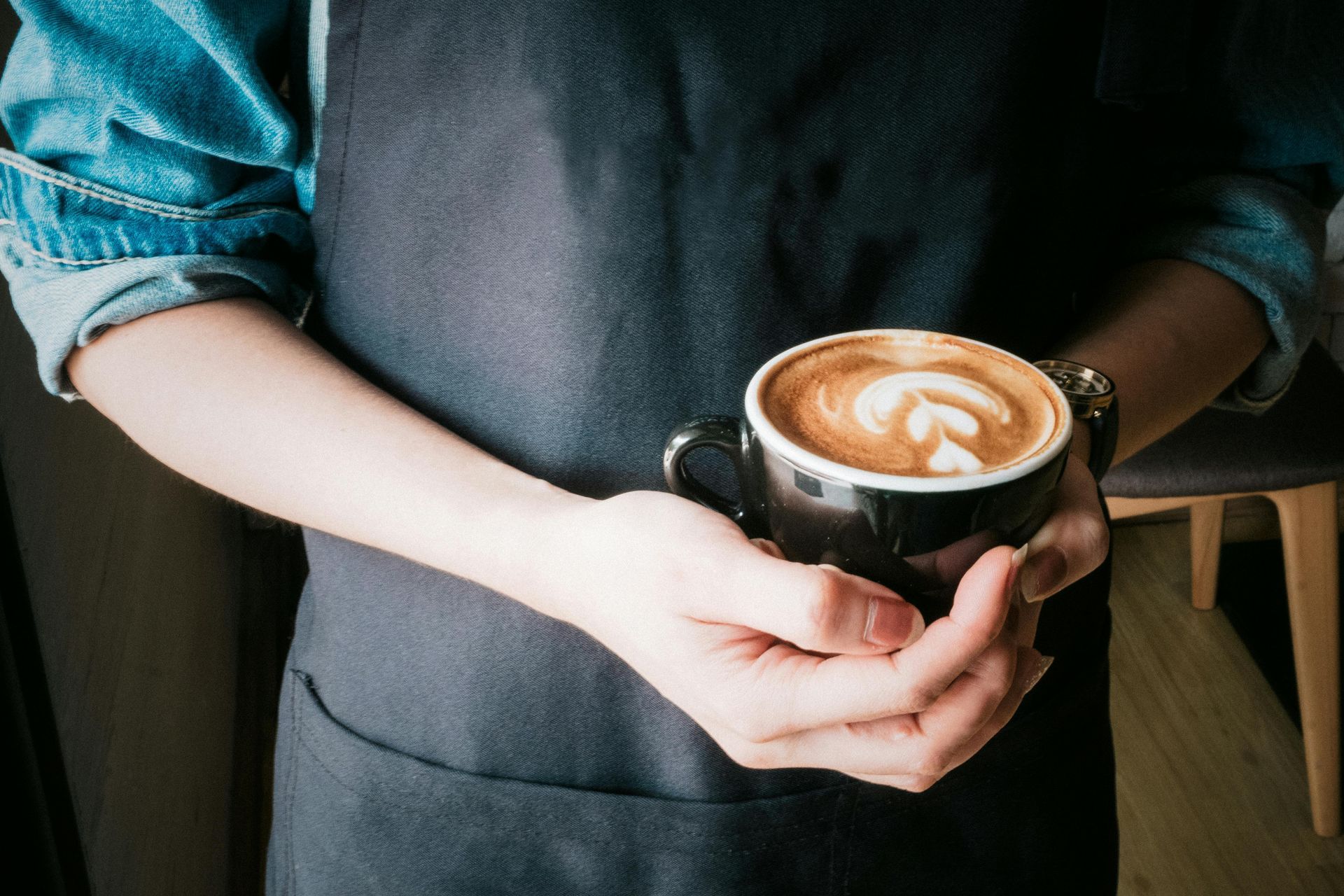 Barista holding a black mug of latte with foam art, wearing a denim shirt and apron.