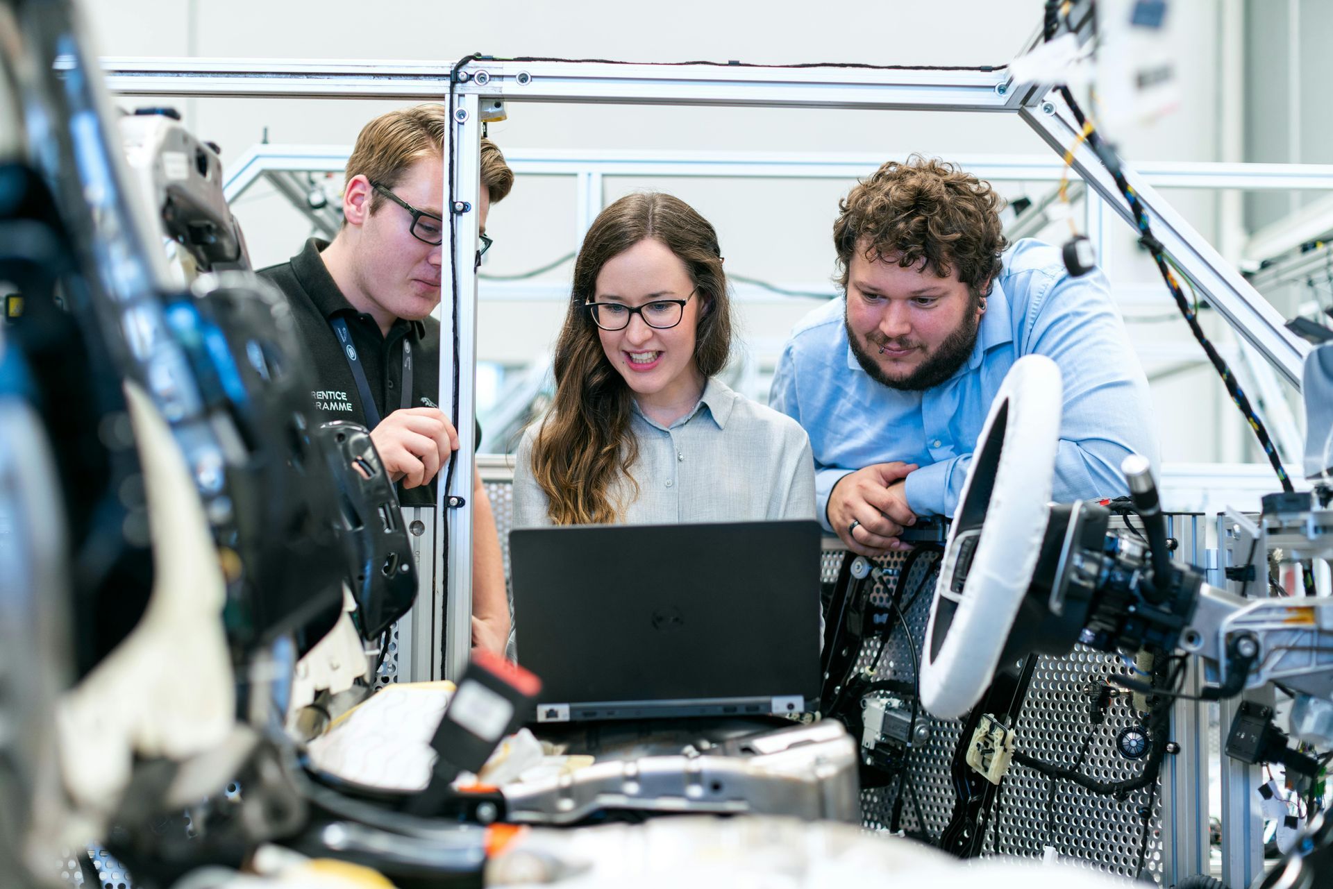 Four engineers examining a laptop near a car chassis in a bright workshop; they appear focused and collaborative.