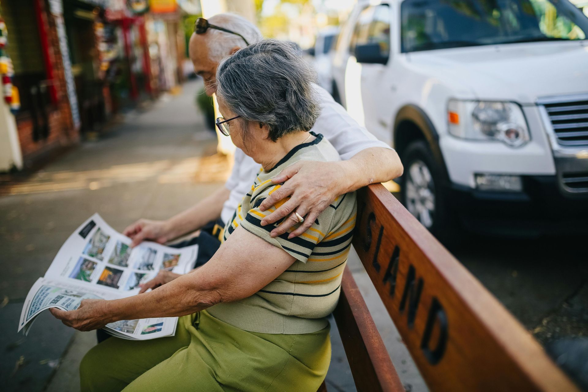 Elderly couple reading a newspaper on a wooden bench, one arm around the other, near a street with a white car.