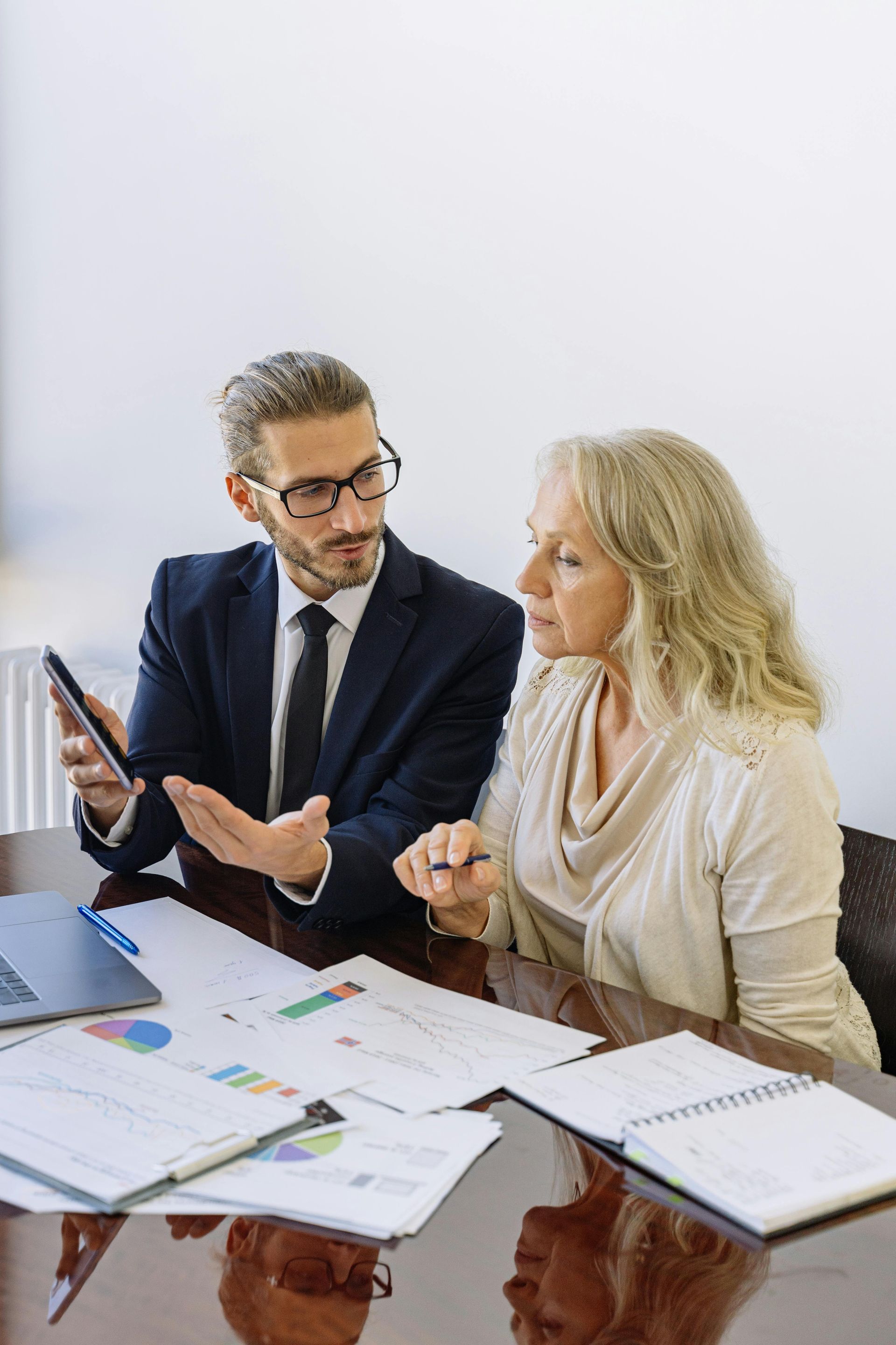 Two coworkers in business attire reviewing charts at a desk, one holding a tablet and the other taking notes.