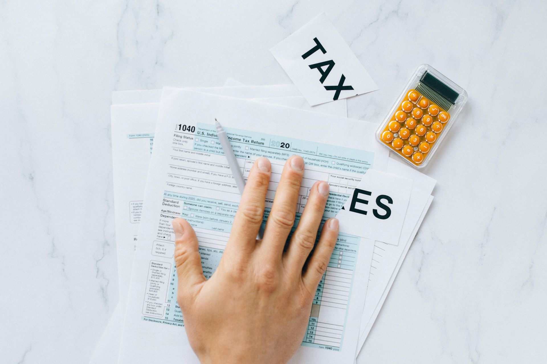 Hand resting on tax forms beside a calculator and pen on a white desk