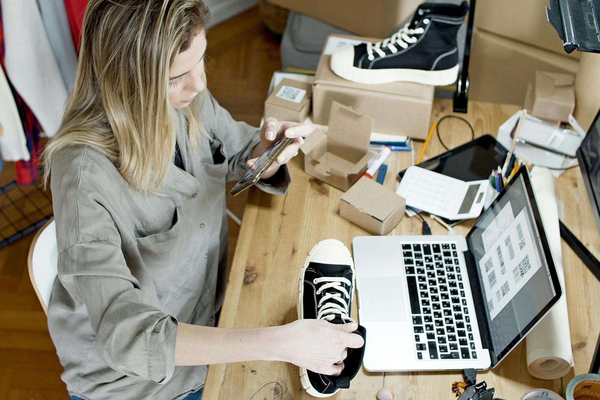 Woman at desk with laptop, photographing a shoe for online sale, surrounded by boxes and other shoes.