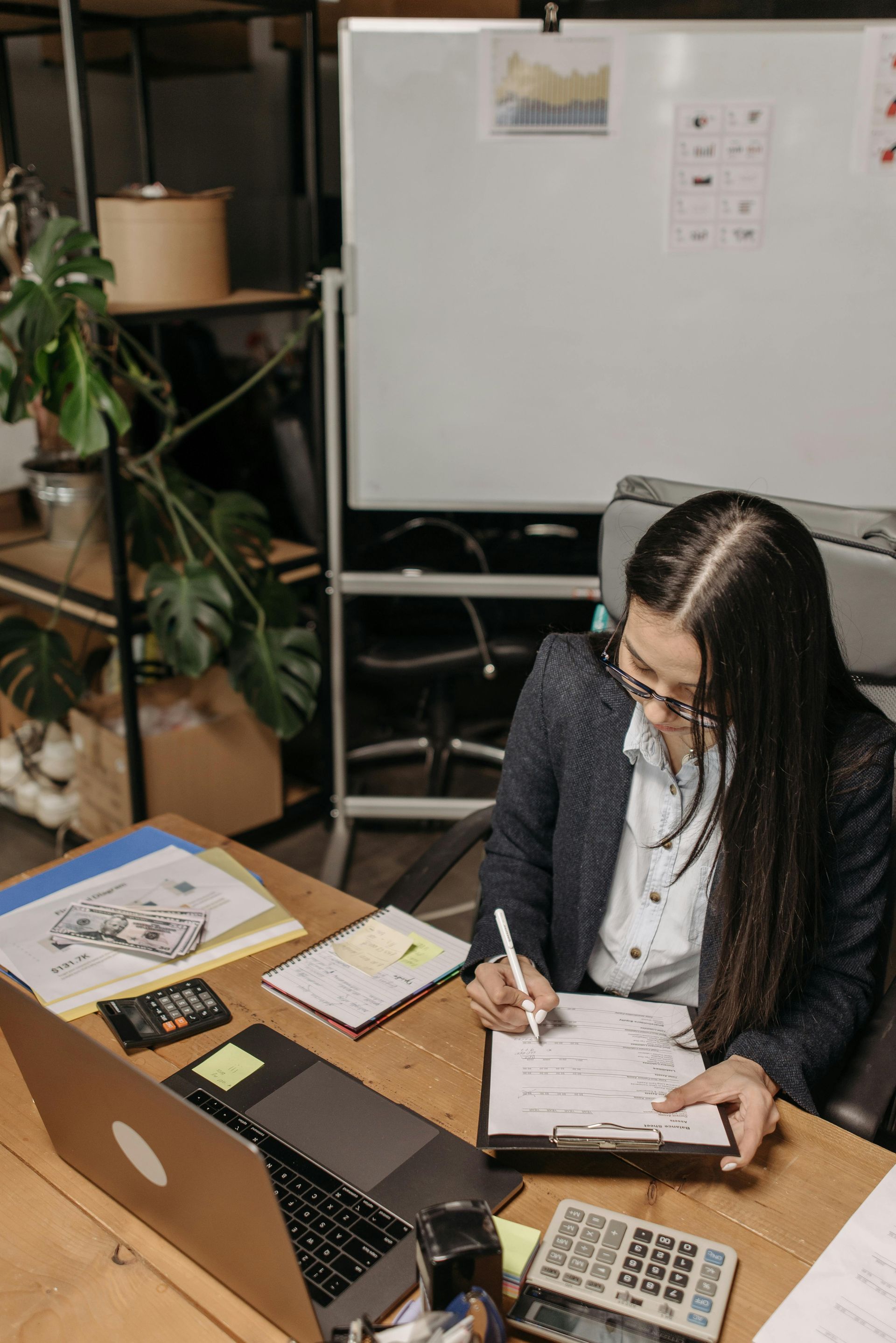 Woman working at a desk, writing on paper beside a laptop in a small office.
