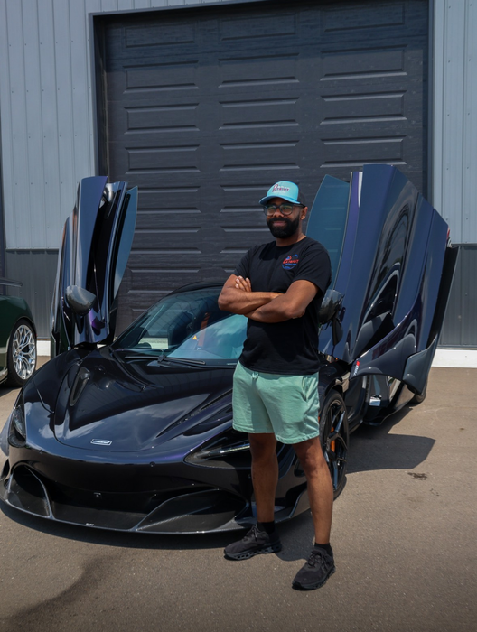 A man with a beard and a hat is smiling in front of a red car.