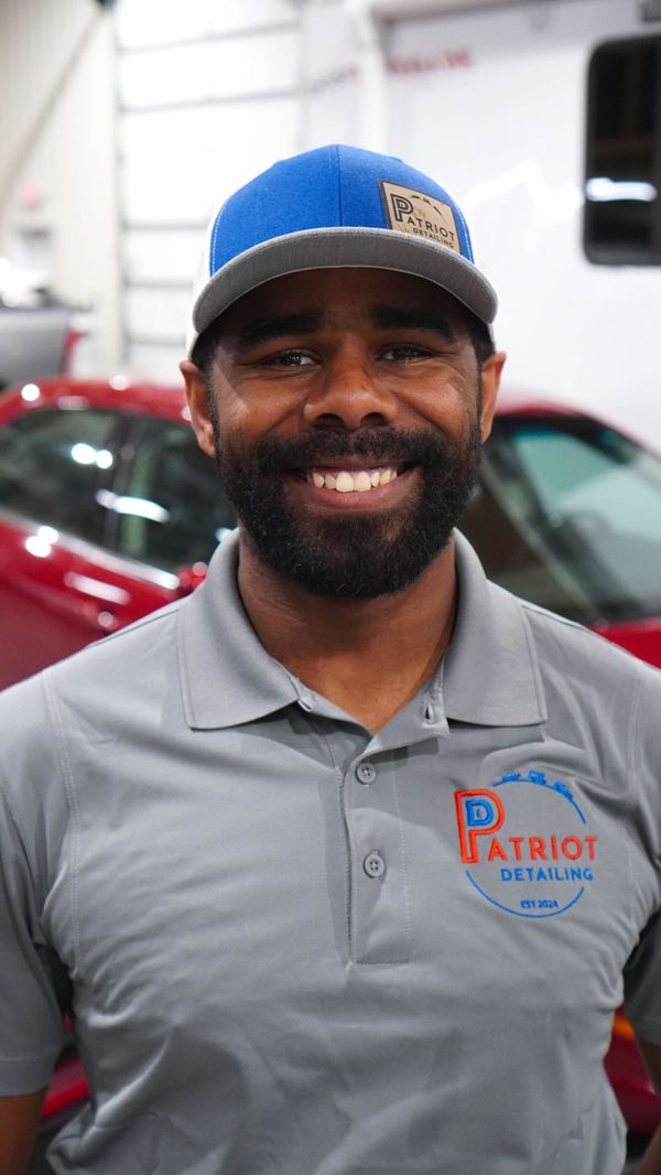 A man with a beard and a hat is smiling in front of a red car.