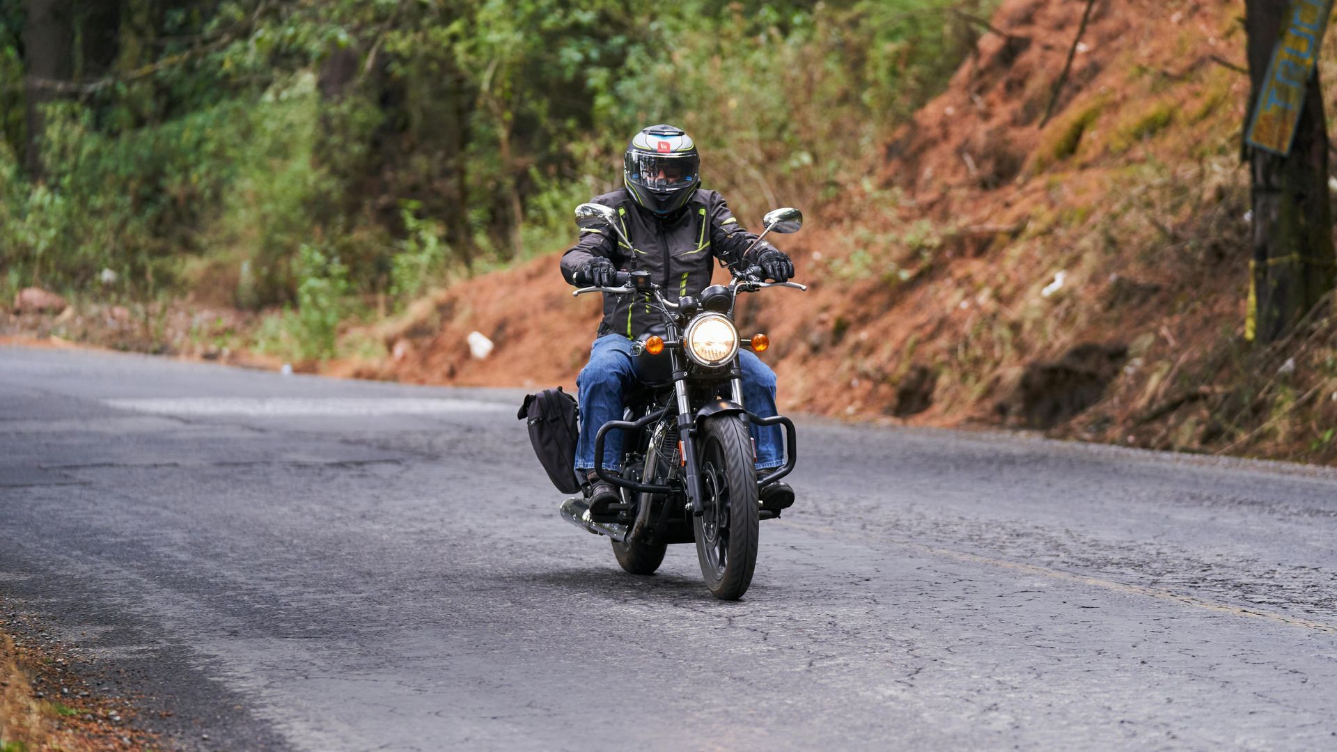 A person in riding gear on a black motorcycle travels along a paved road flanked by a wooded, earthen embankment.