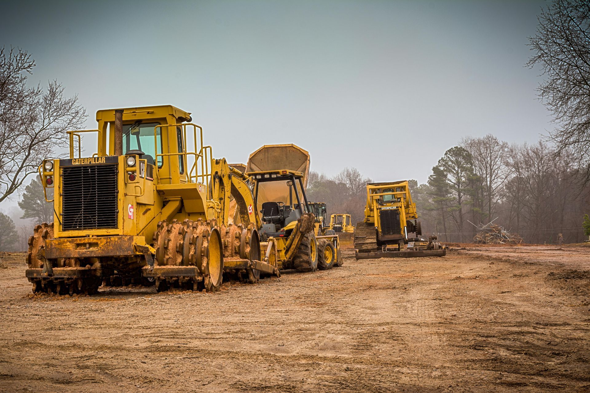 Three yellow construction vehicles sitting on a dusty dirt field under an overcast sky.