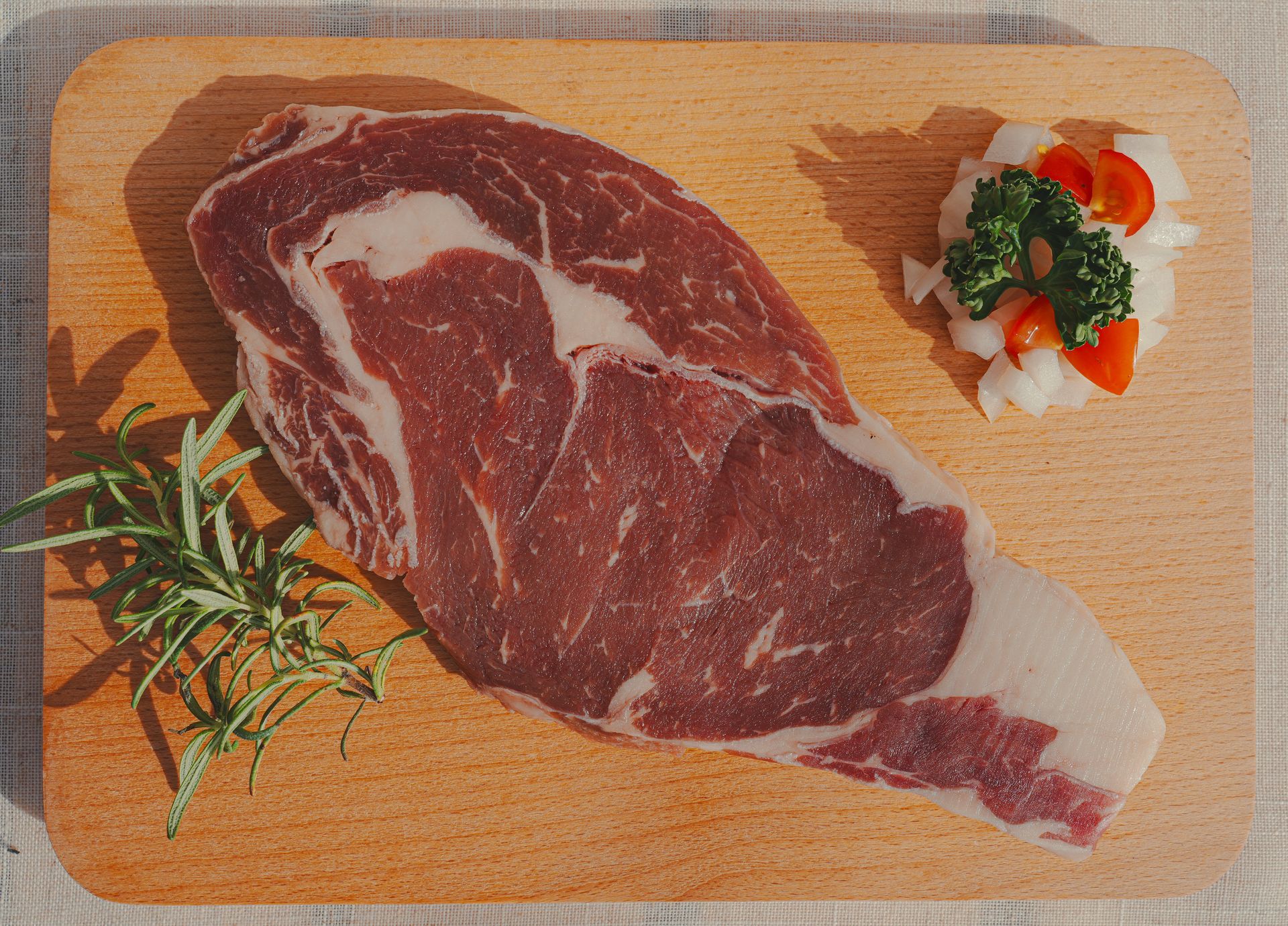 一塊帶有大理石紋理的生肋眼牛排放在木質砧板上，配上迷迭香、洋蔥和番茄。