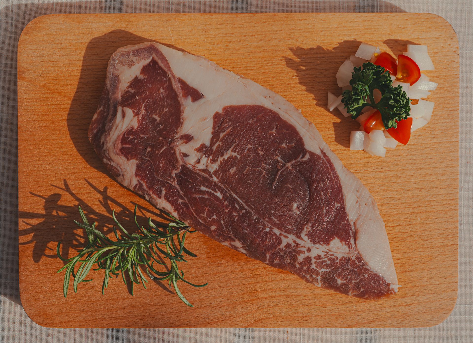 一塊帶有脂肪紋理的生牛排，配上香草和切碎的蔬菜，放在木製砧板上。