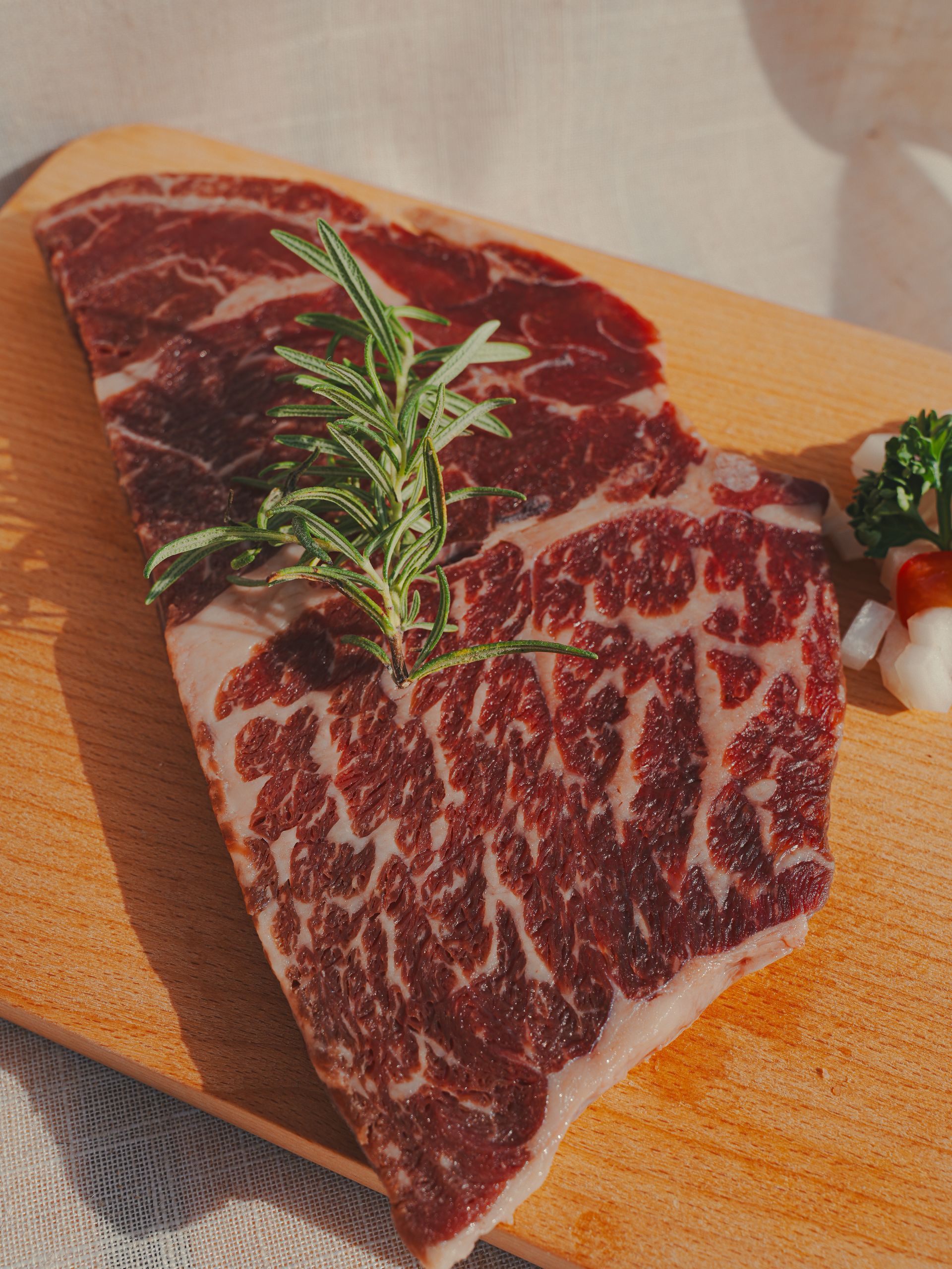 一塊帶有大理石紋理的生牛排，配上迷迭香枝和一些蔬菜，放在木板上。