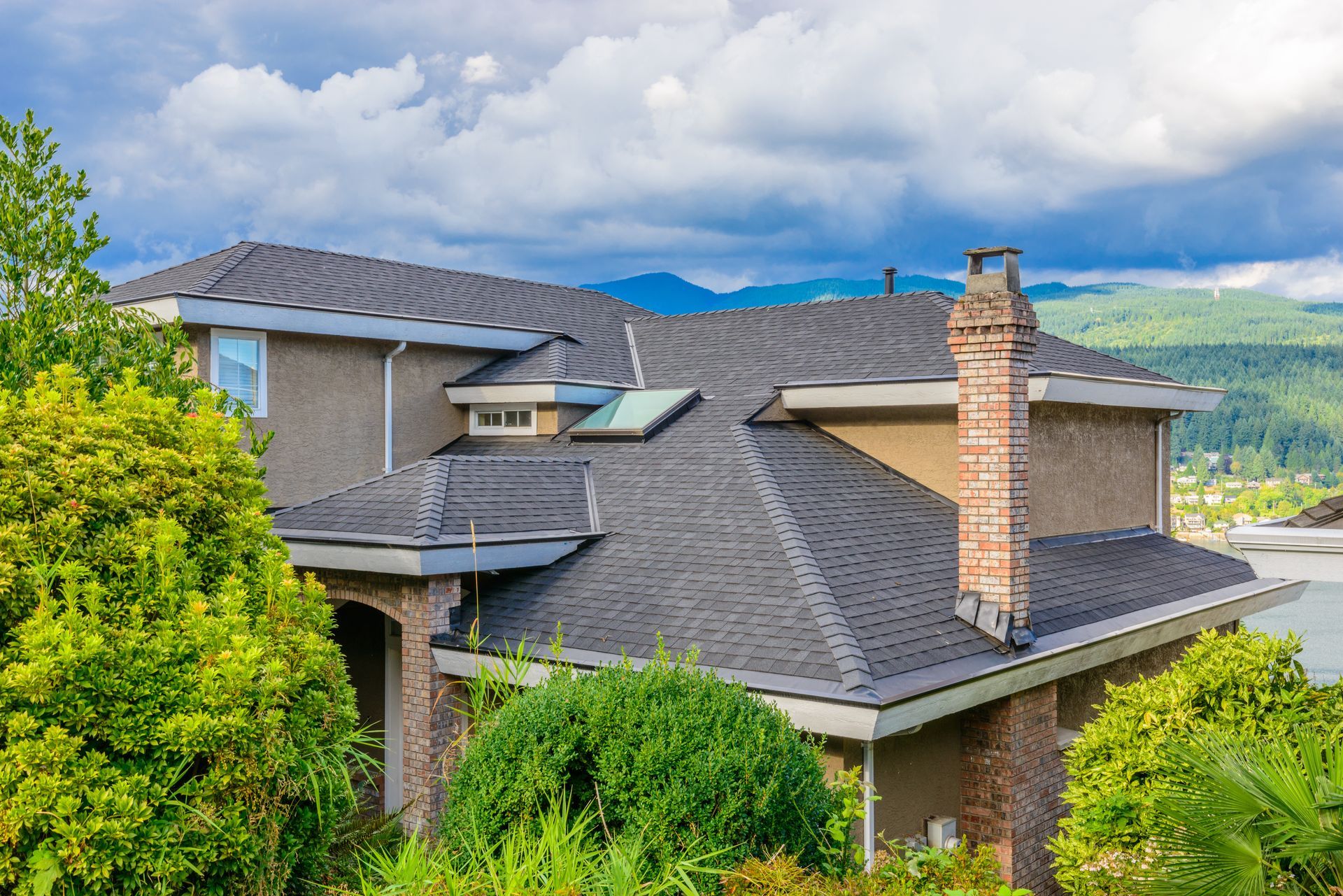 Two-story beige house with a dark gray shingled roof, chimney, and surrounding green trees against a cloudy sky.