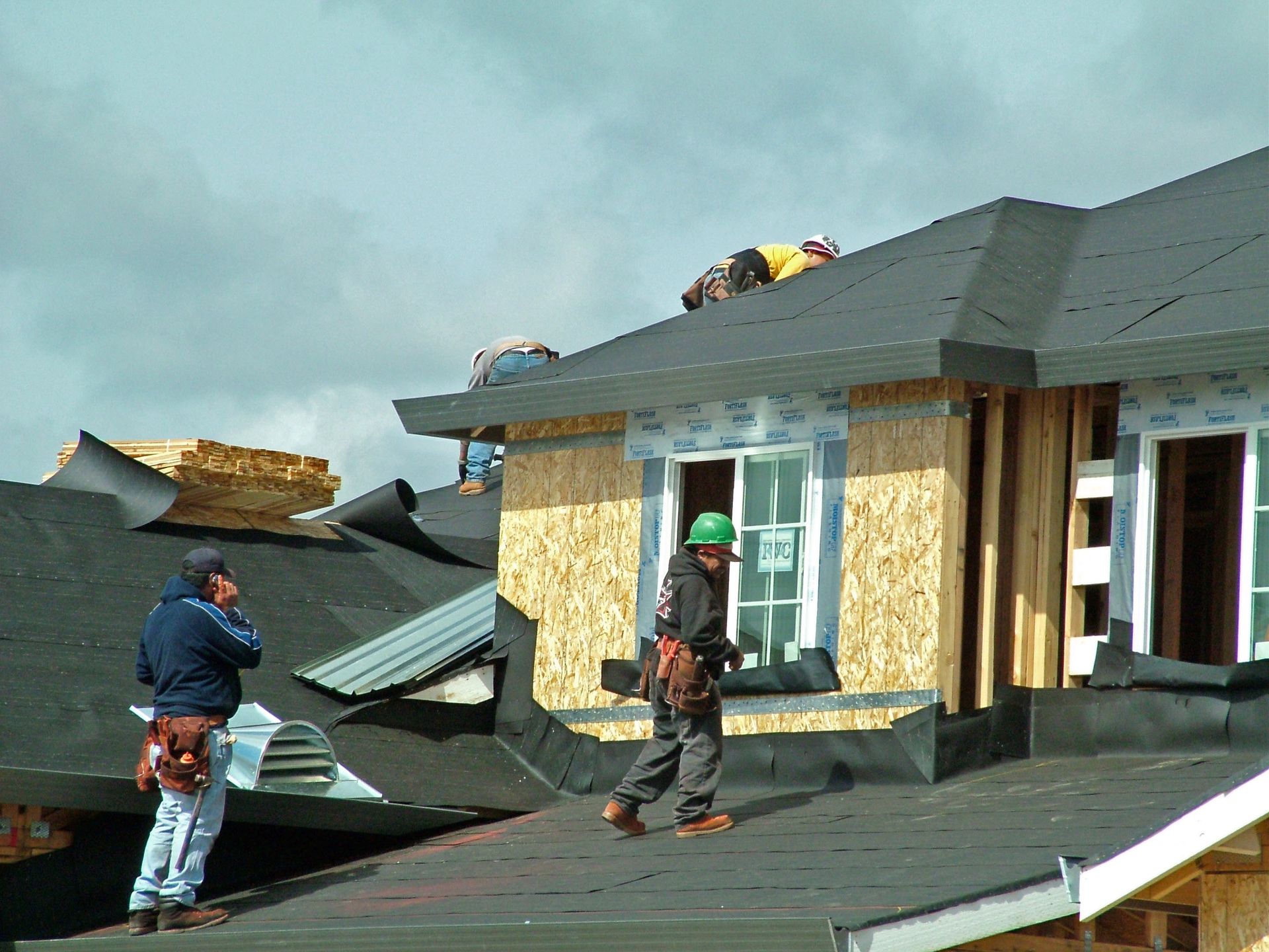 Construction workers on a roof, installing shingles on a new building under a cloudy sky.