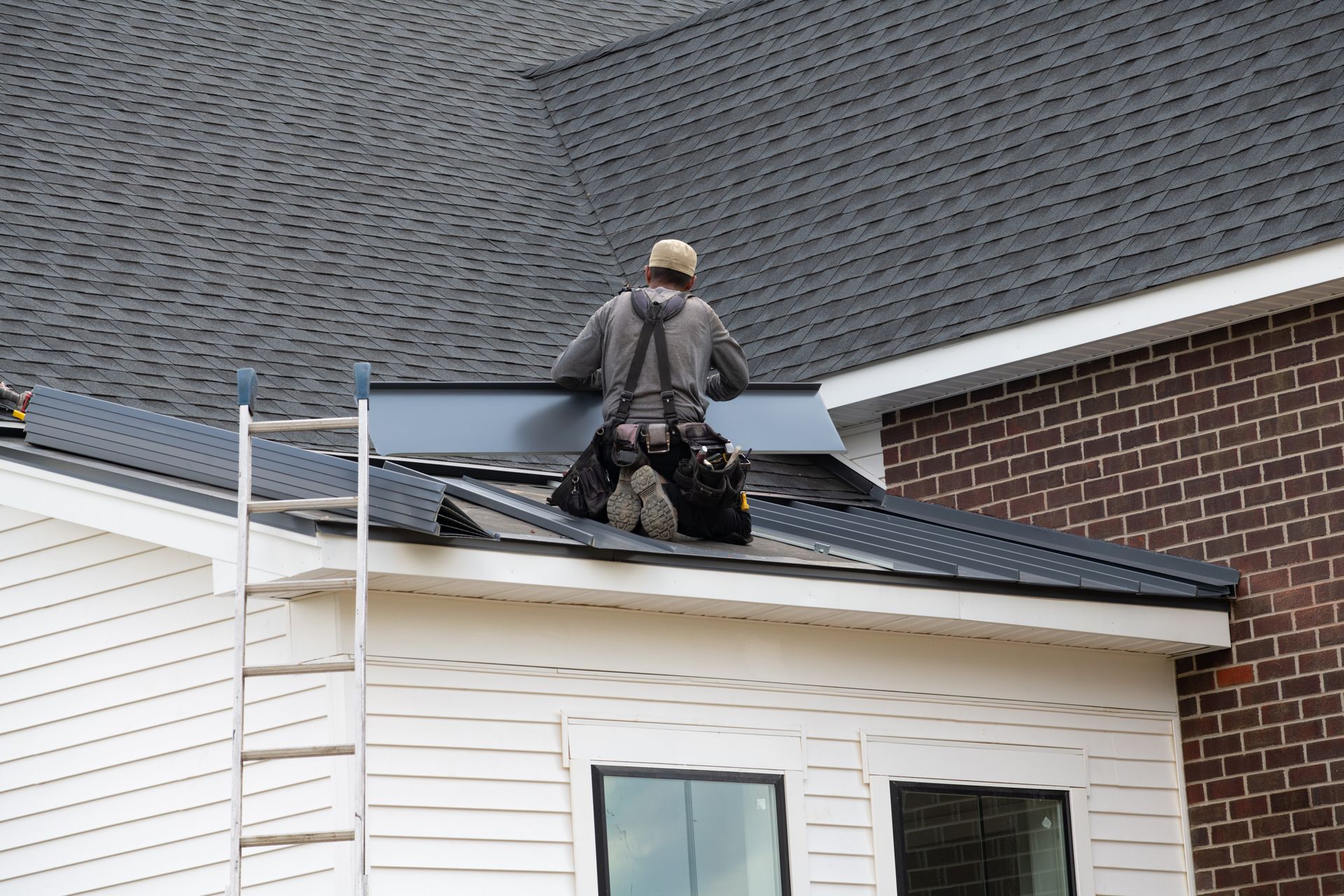 Roofer installing dark metal roofing, kneeling on a roof with a ladder nearby.