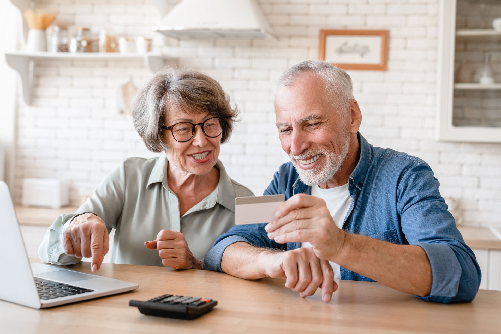 Older adults using a laptop, one holding a credit card, smiling in a kitchen.