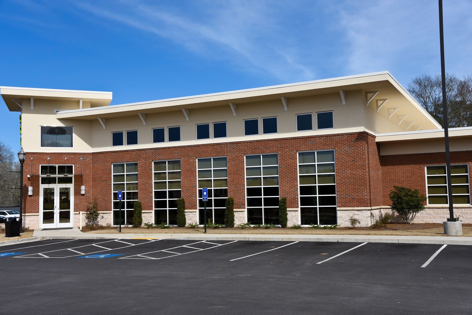 Brick building with large windows and a sloped roof under a blue sky; parking lot in front.