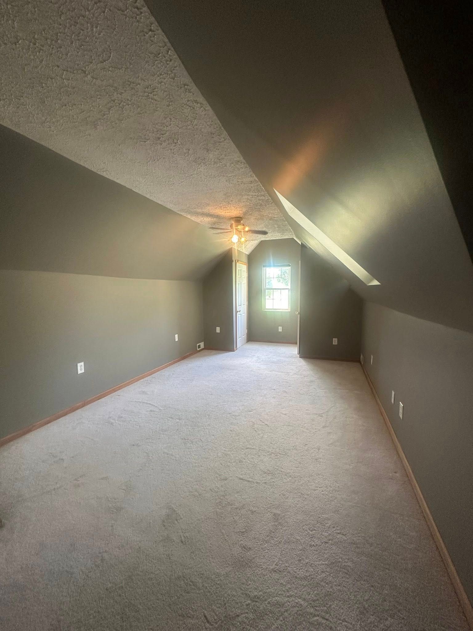 Empty room with sloped ceilings, gray walls, carpet. Window at the end of the room.
