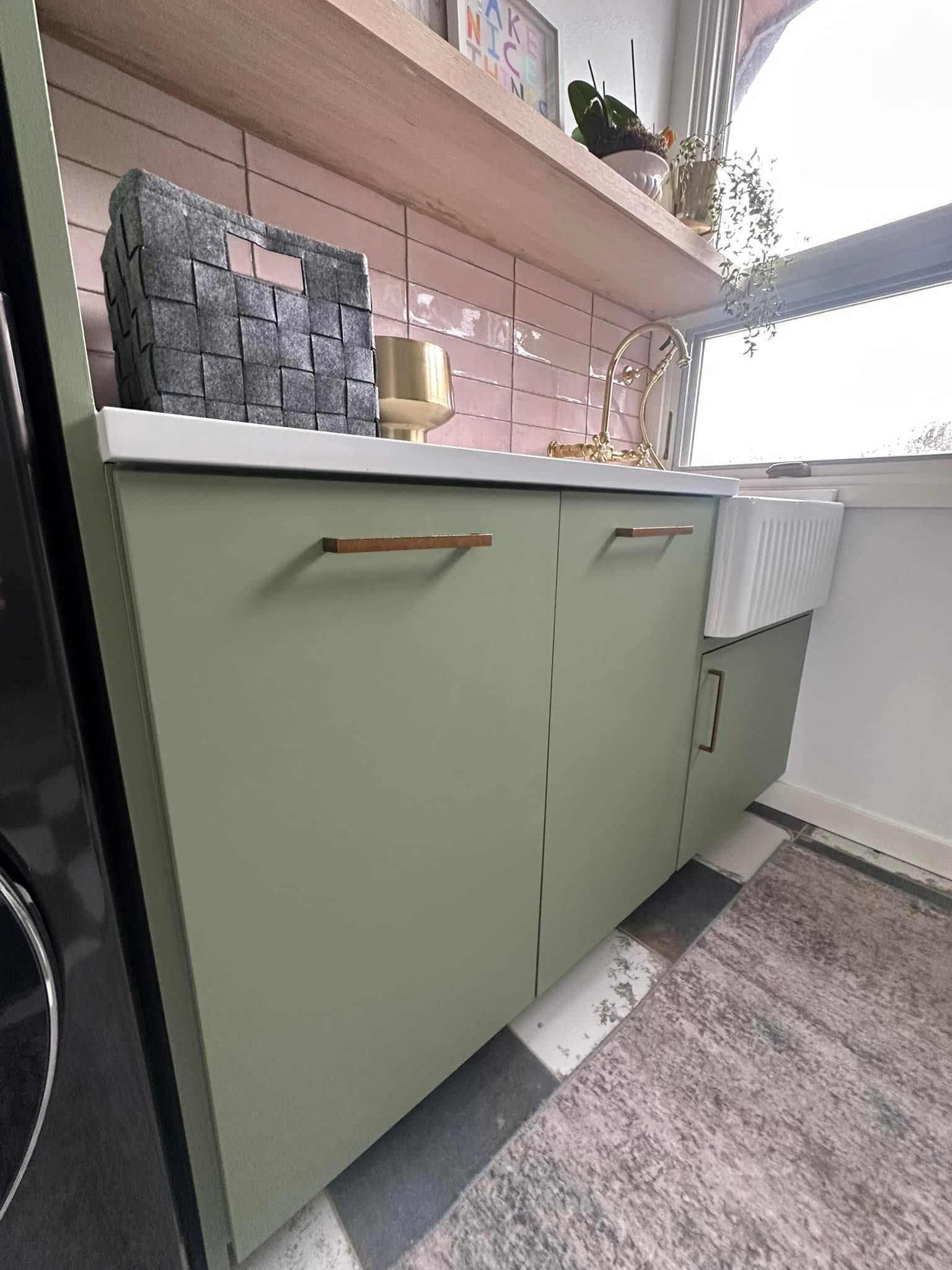 Green cabinets with wooden handles, a white countertop, and a shelf with decor in a laundry room.