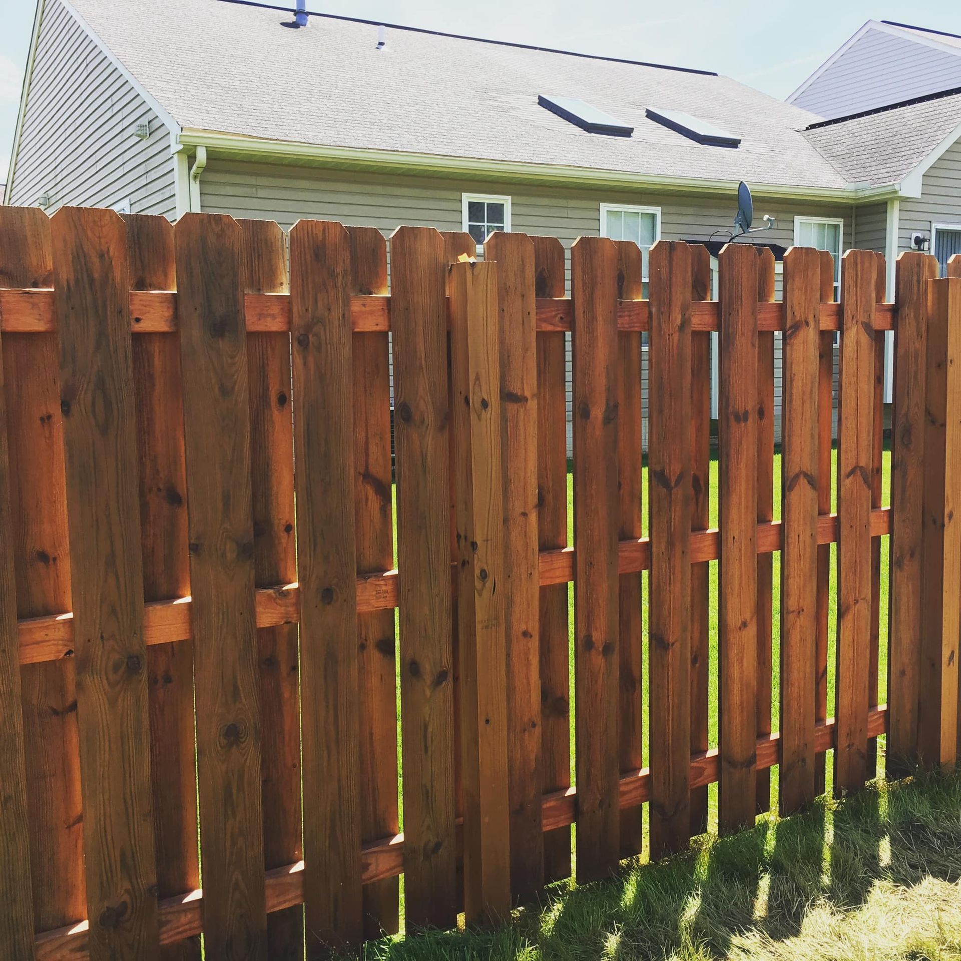 Wooden picket fence with brown stain, in front of a light gray house with grass and sunlight.