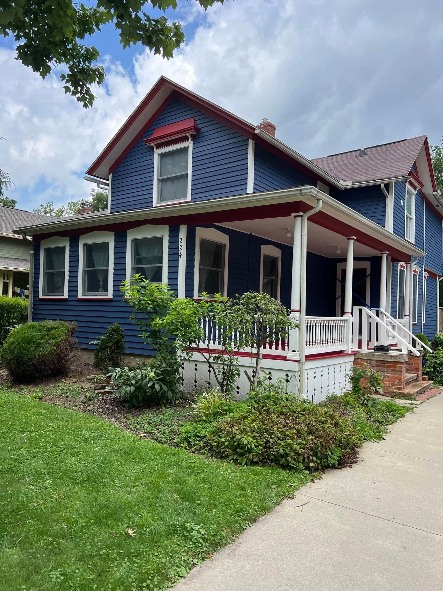 Blue house with white trim, red accents, and a porch surrounded by greenery on a sunny day.