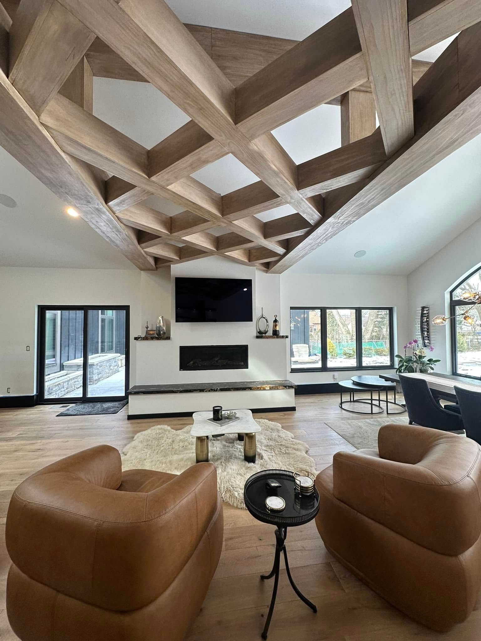 Spacious living room with wood-beamed ceiling, fireplace, leather armchairs, and large windows overlooking the outdoors.