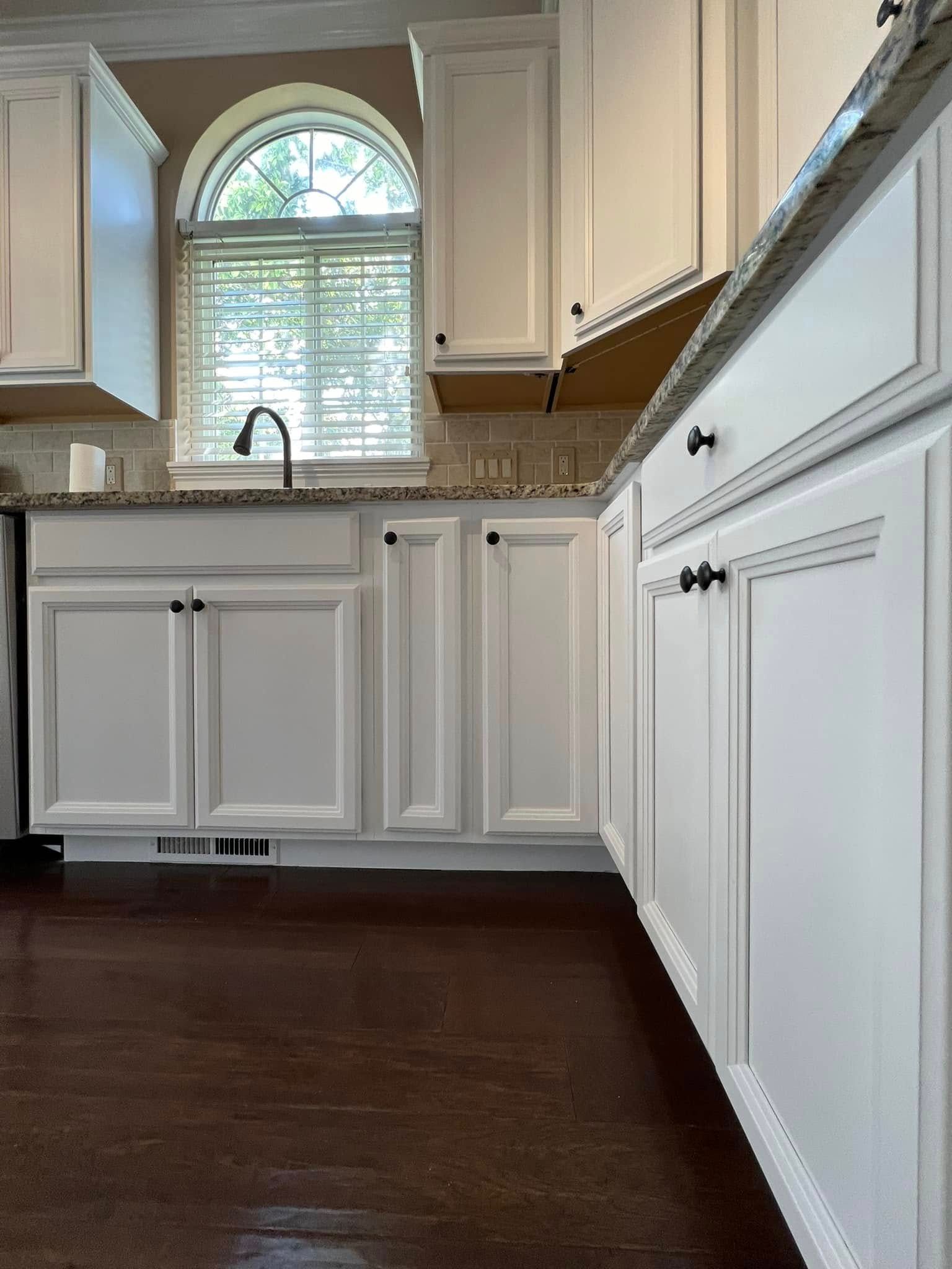 White kitchen cabinets with black hardware, dark wood floor, and arched window with blinds.
