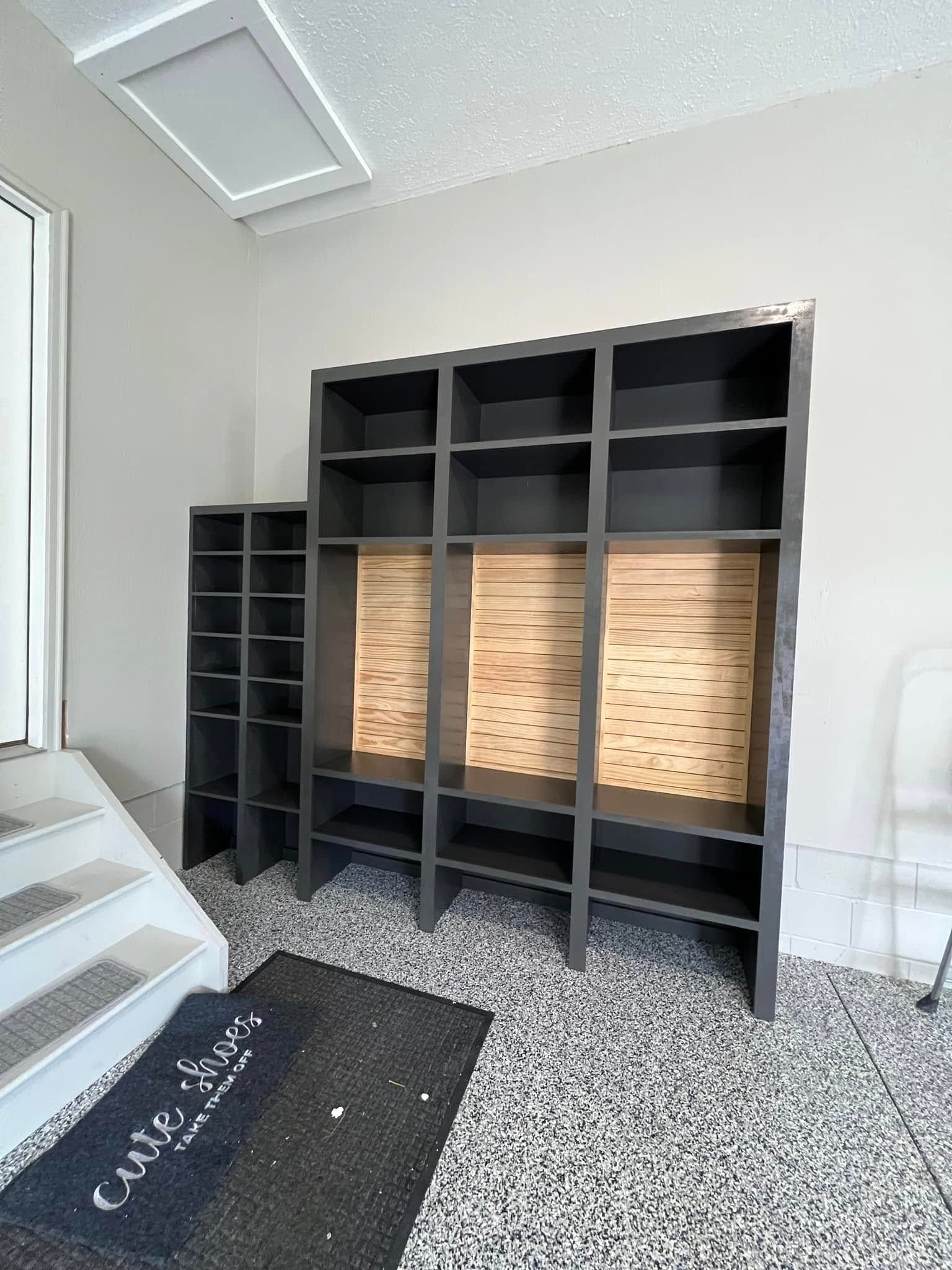 Dark gray built-in shelving unit with wood backing, next to a narrow shoe rack.  Mat on speckled floor.