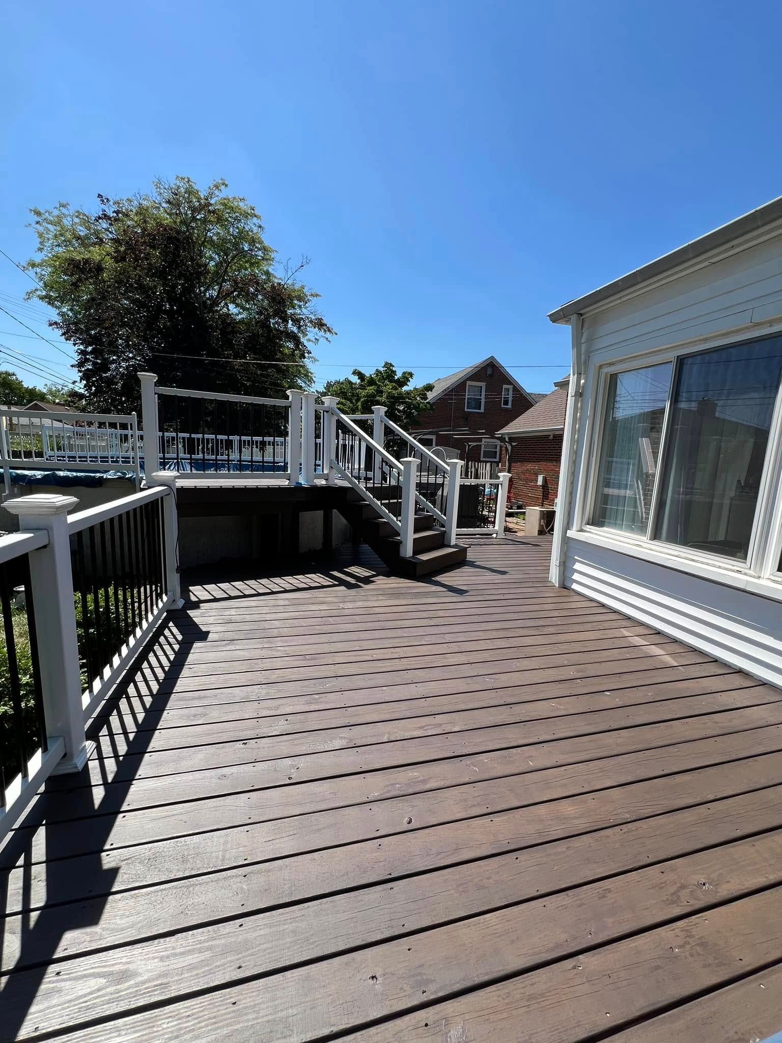 Wooden deck with stairs and white railing, house with windows in the background, clear blue sky.