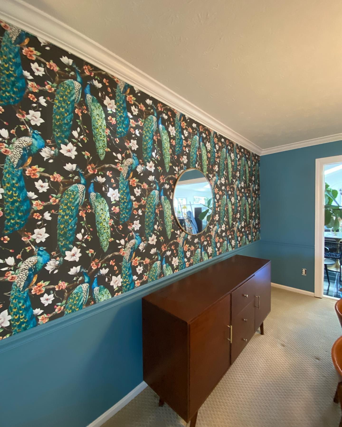 Wall with peacock wallpaper, blue paint, wooden cabinet, and oval mirror.