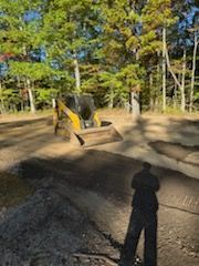 Yellow skid steer operating on a dirt surface, with trees in the background and a person's shadow in the foreground.
