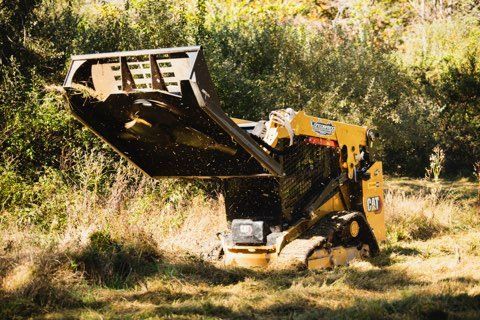 Yellow track loader with raised bucket, mulching grass in a field.