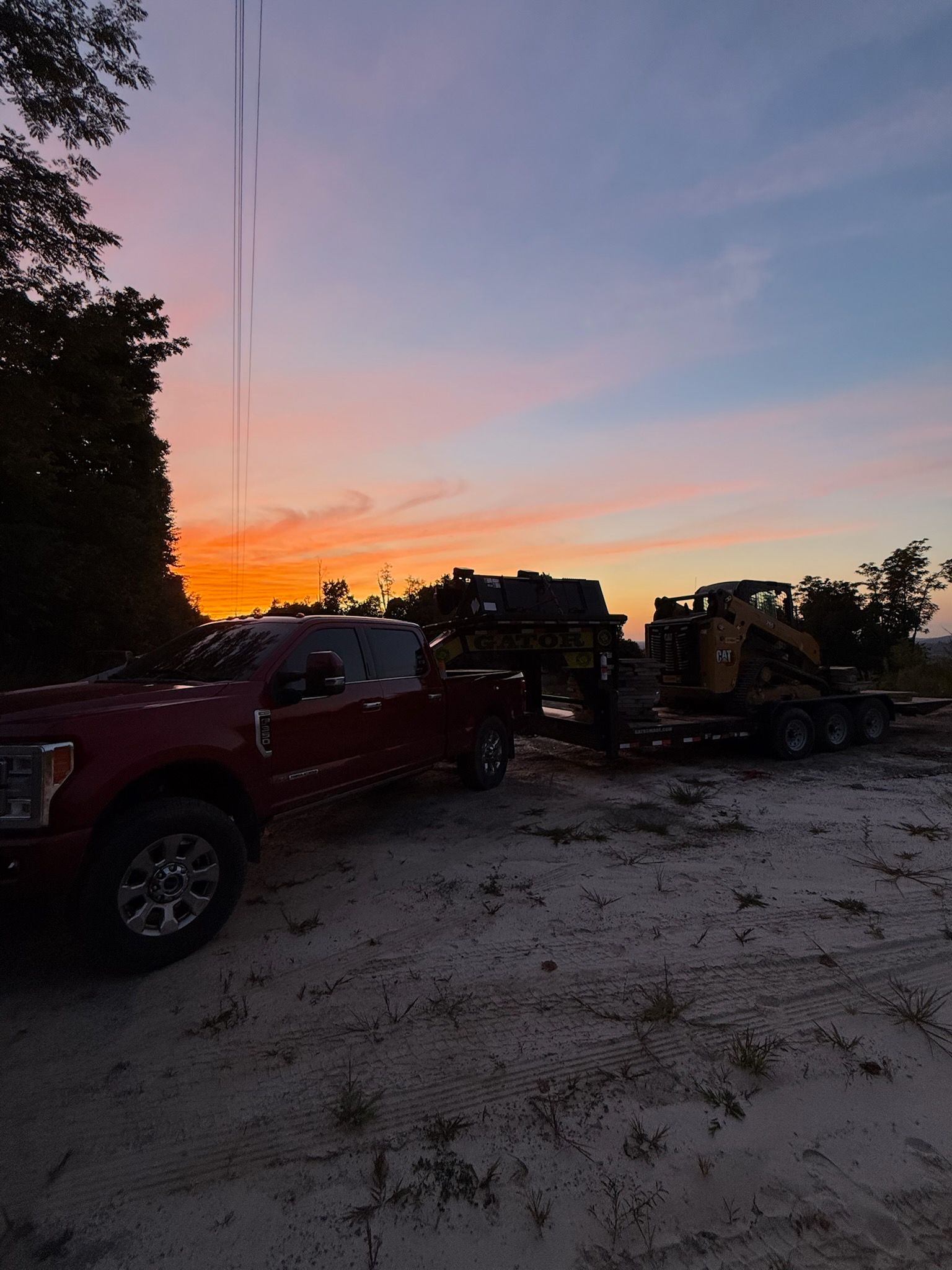Red pickup truck towing equipment on a trailer at sunset with orange and blue sky.