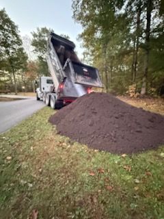 Dump truck unloading a large pile of dark brown mulch onto a grassy area next to a road, autumn setting.